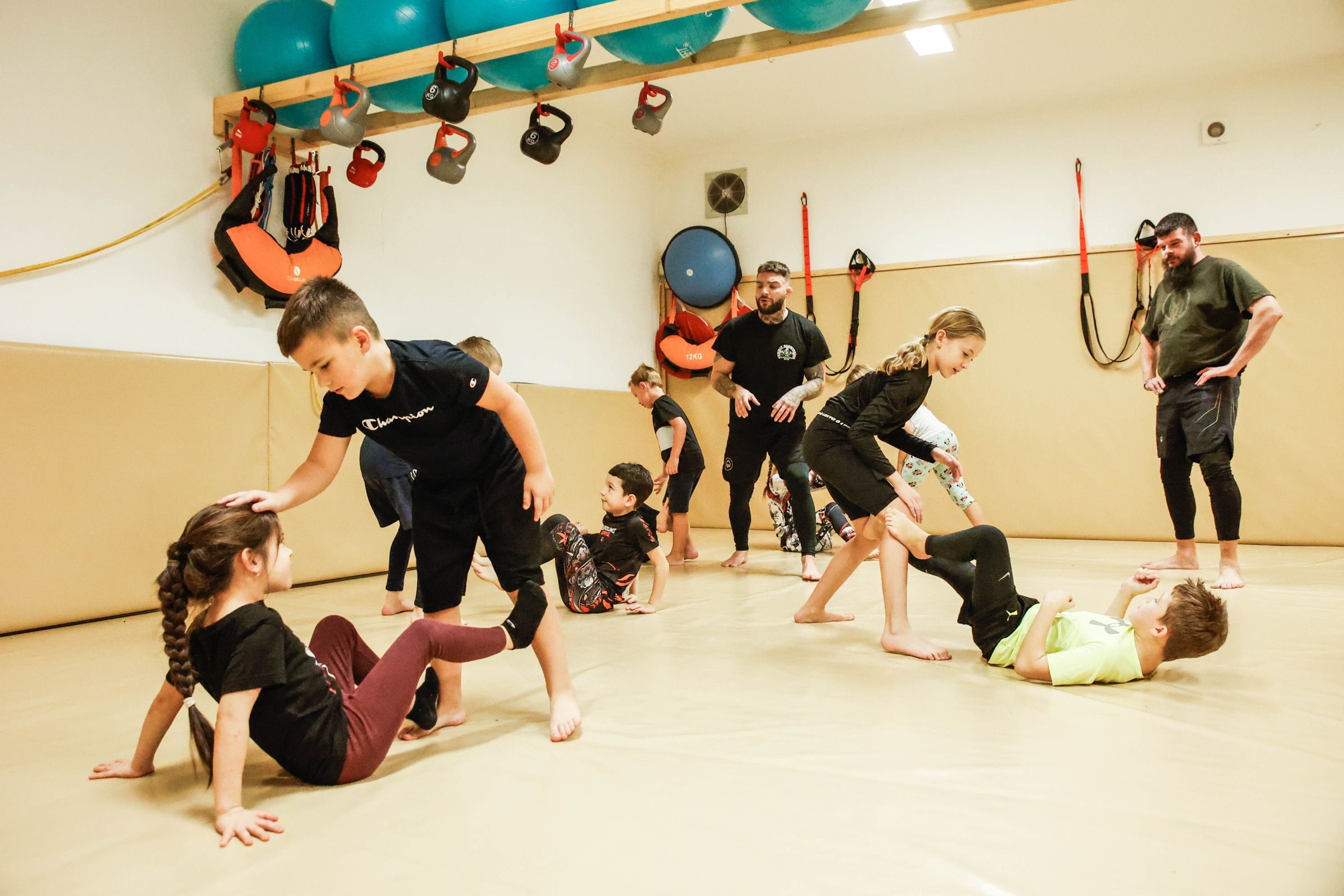 Děti nacvičují ve dvojicích na tatami, trenér stojí mezi nimi a sleduje je.